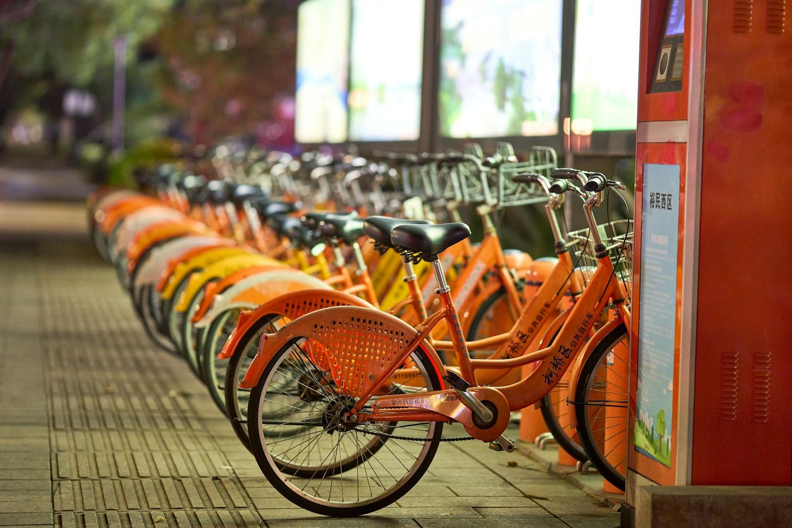 Lång rad av orangea hyrcyklar parkerade utomhus på en trottoar kvällstid.